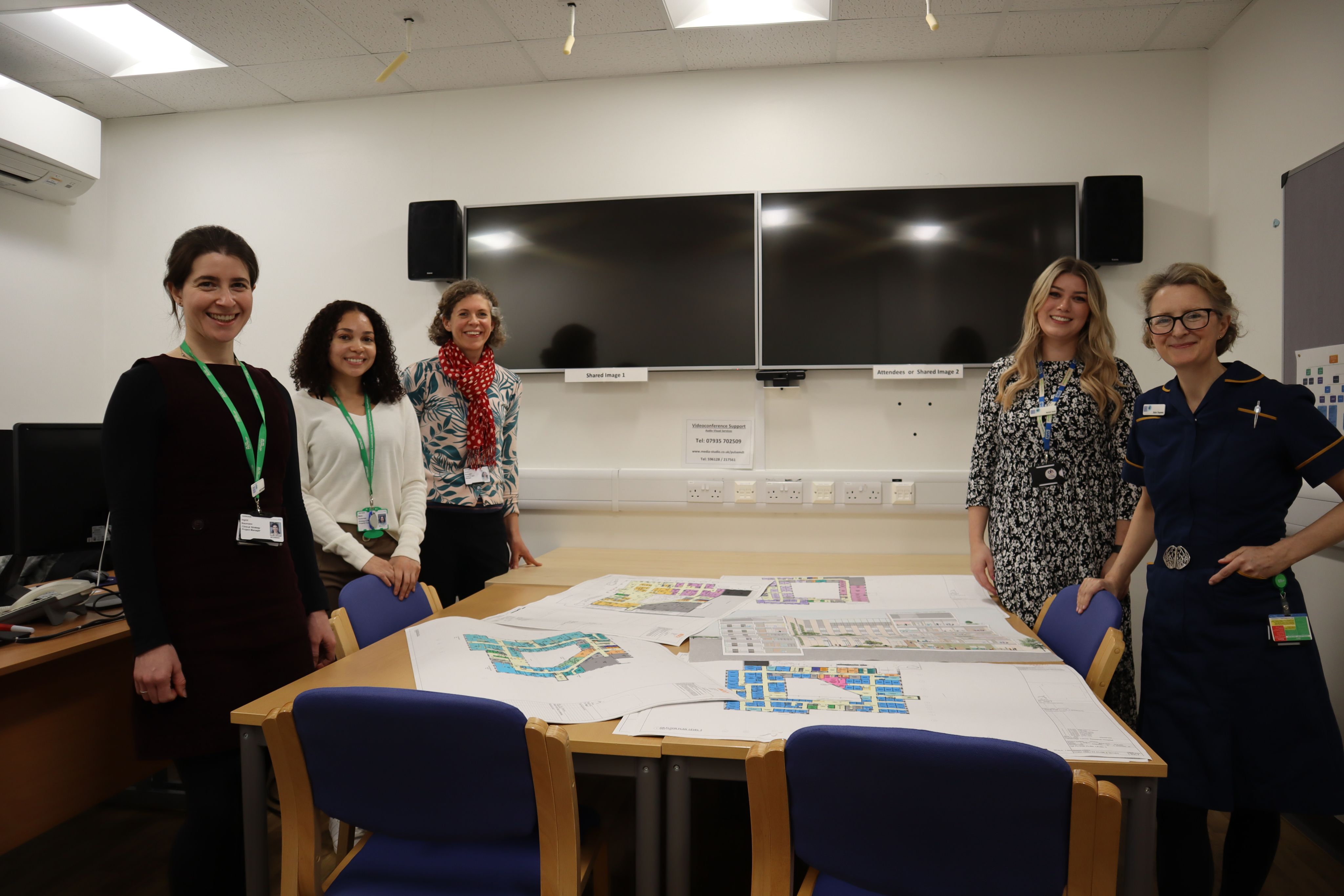 Five members of the team are standing round a table with the floorpans on for the new hospital. The people are looking at the camera and smiling.